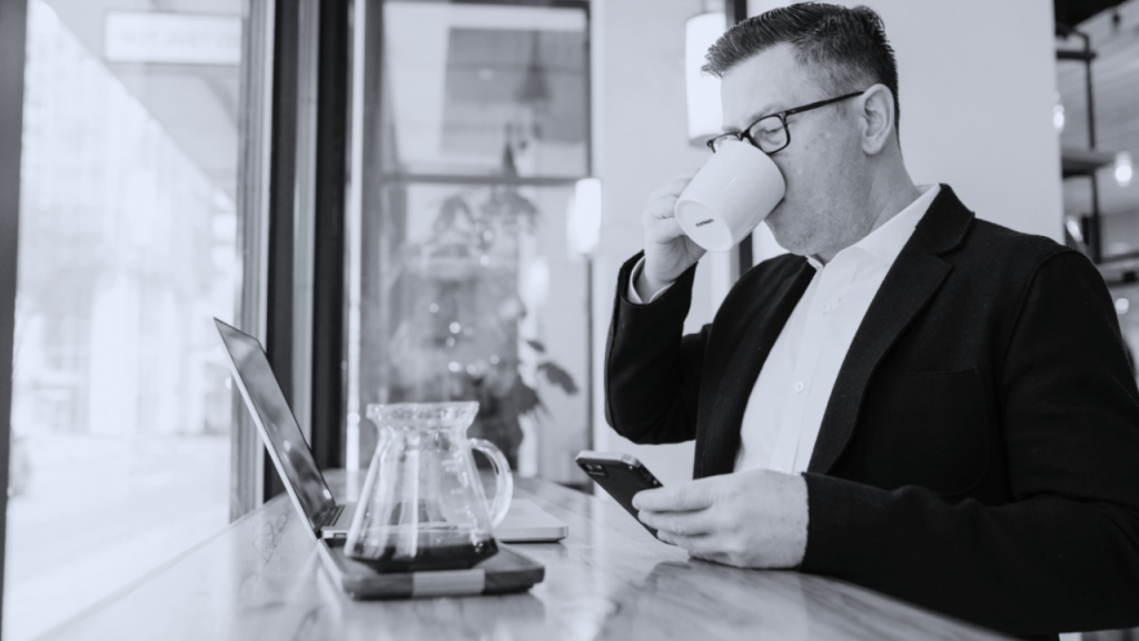 consultor de vendas, sentado em um café tomando algo em uma xicará olhando o celular e notebook